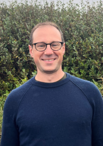 Headshot of attorney David Woodsmall. He has short brown hair and glasses and is wearing a navy sweater while grinning at the camera.