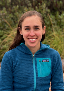 Headshot of climate attorney Morgan. She has brown hair and a bright smile and is wearing a turquoise blue jacket in front of a grassy background.
